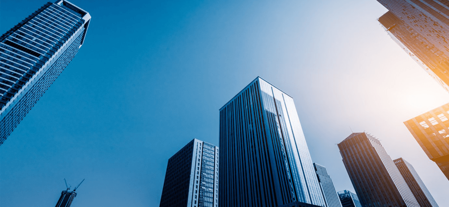 Looking up at tall office buildings with blue sky and sun visible on right side.