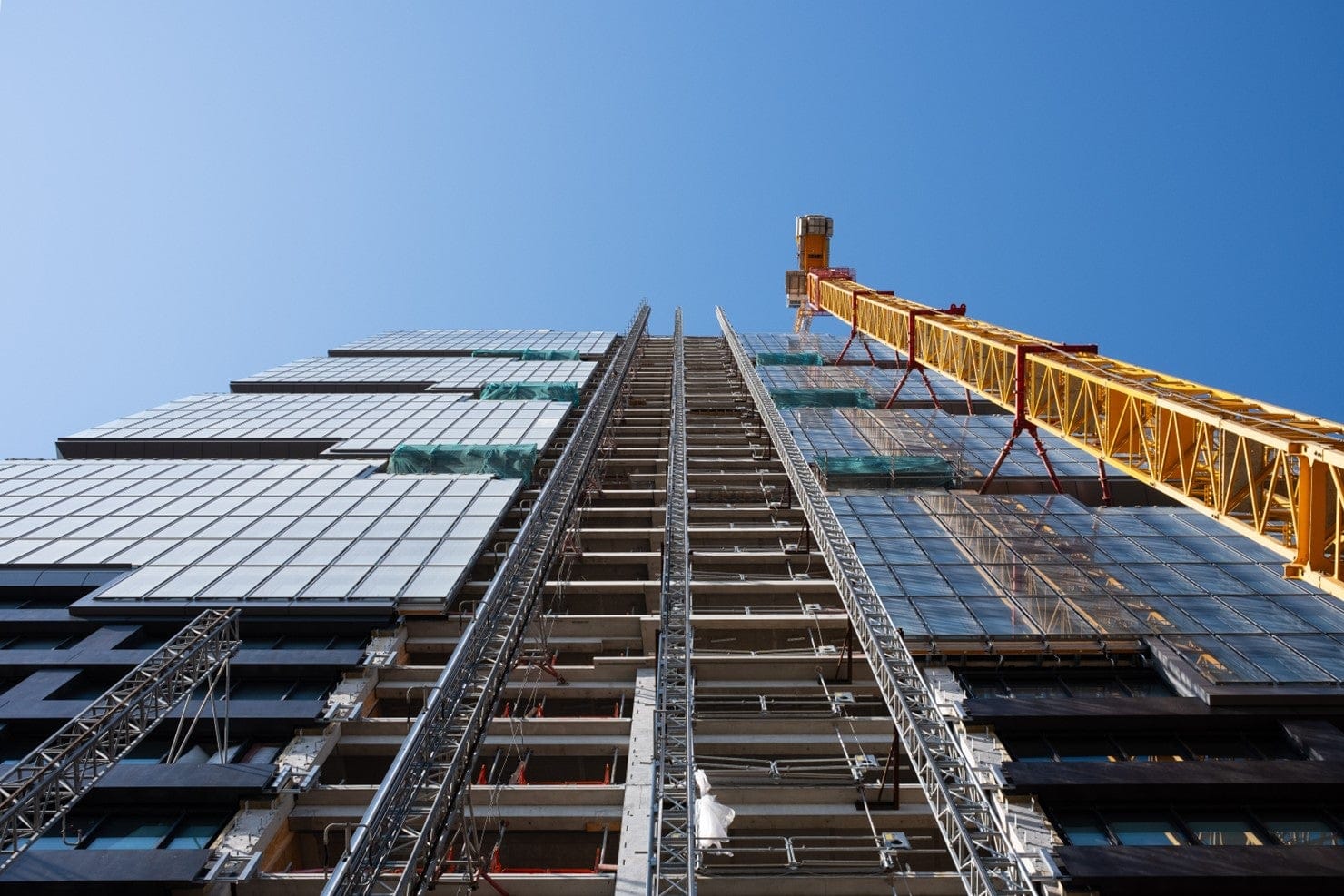 View from ground looking up at tall office building being constructed, with yellow crane attached to building on right side.